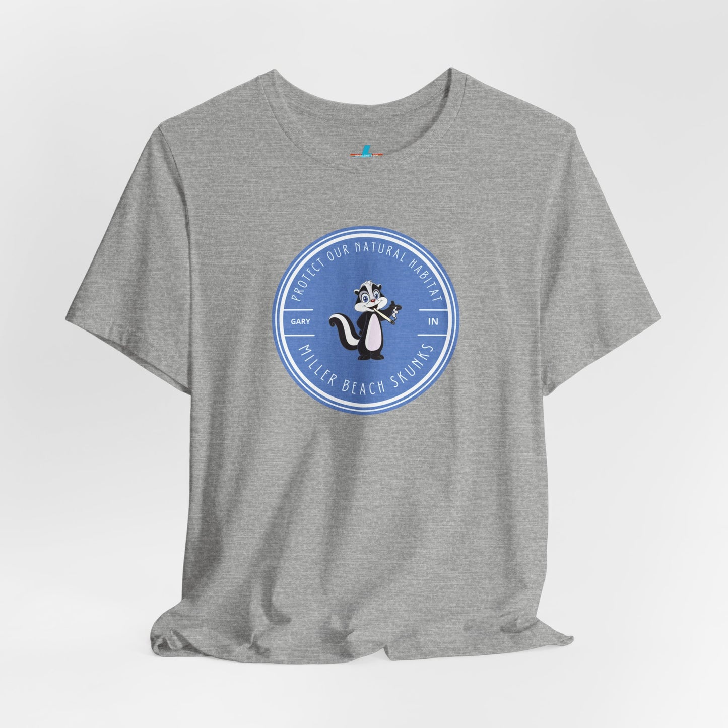 The Printify Miller Beach Skunks - Unisex Jersey Short Sleeve Tee is a gray T-shirt that features a circular blue logo at the center. Inside the logo, there is an illustration of a skunk with the text "Protect Our Habitat" and "Miller Beach Skunks" around the border, promoting environmental consciousness. The upper left part of the logo reads "Gary, IN.