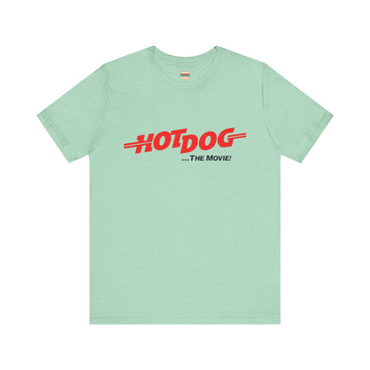 A smiling man and woman interact while wearing matching yellow "Hot Dog The Movie 1984 - Unisex Jersey Short Sleeve Tee" by Printify, featuring bold red "HOT DOG THE MOVIE" text. Their vibrant tees perfectly complement their blue jeans, with the man casually having his hands in his pockets and the woman resting one hand on his shoulder.