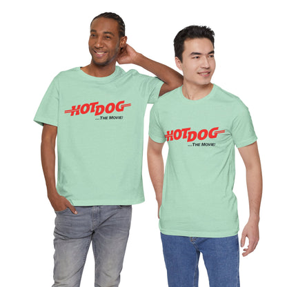 A smiling man and woman interact while wearing matching yellow "Hot Dog The Movie 1984 - Unisex Jersey Short Sleeve Tee" by Printify, featuring bold red "HOT DOG THE MOVIE" text. Their vibrant tees perfectly complement their blue jeans, with the man casually having his hands in his pockets and the woman resting one hand on his shoulder.