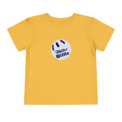 A young child with light hair stands against a plain white background, smiling while wearing a vibrant yellow Printify short sleeve tee named "Miller Wiffel Ball OG Logo - Toddler Short Sleeve Tee." The shirt features the "Miller Wiffle" text and a wiffle ball graphic on the front. The child is also dressed in blue pants.