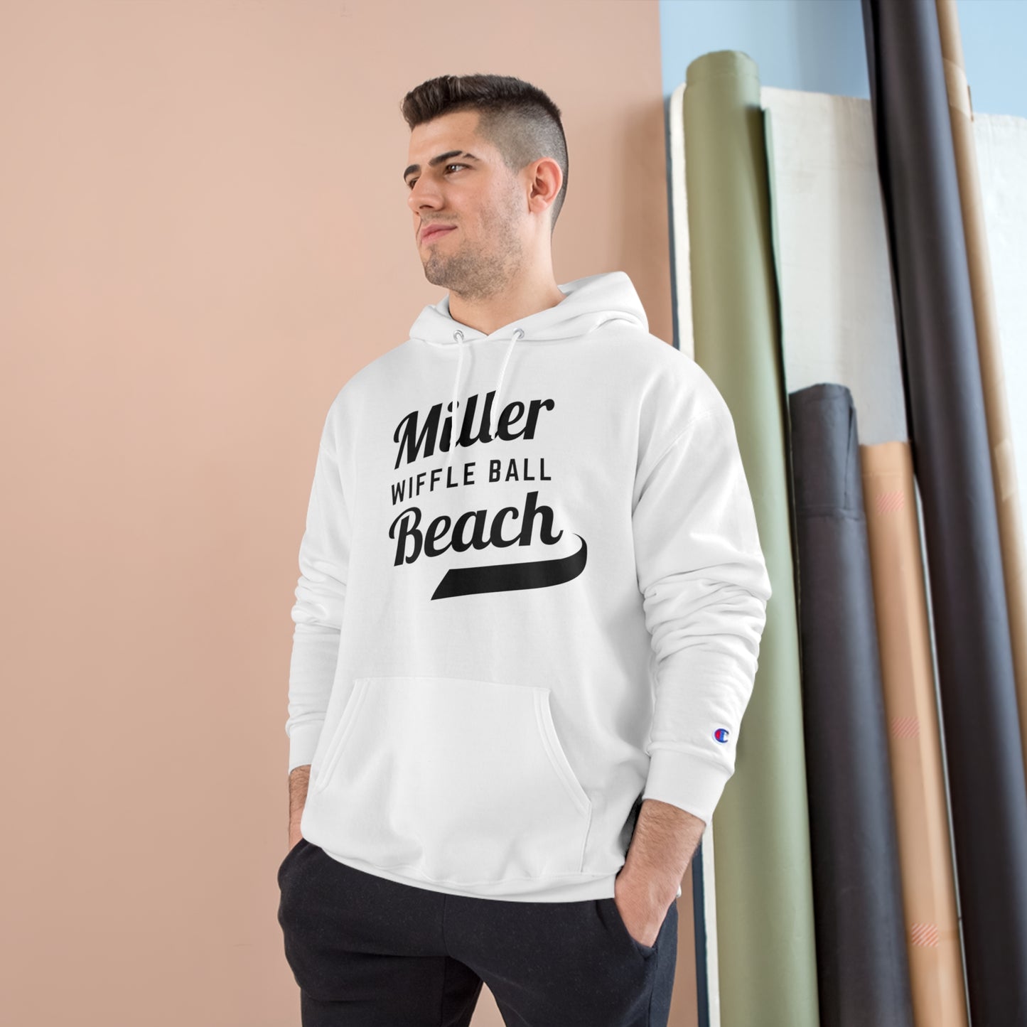 A man wearing a Champion Hoodie from Printify, featuring the "Miller Beach Wiffel Ball Retro Jersey Logo," stands against a peach-colored wall and vertical rolls of various colored fabrics. He has short, dark hair and is looking slightly to the left with a relaxed expression.