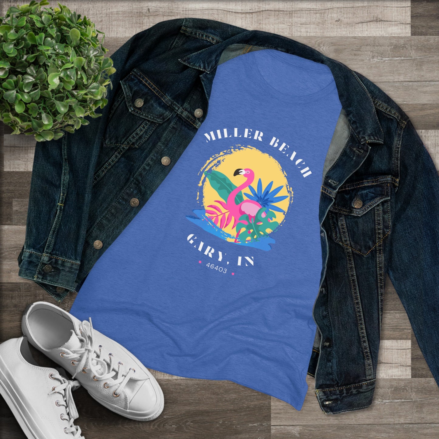 A women's triblend tee from Printify, the Miller Beach Flamingo features a blue color and "Miller Beach Gary, IN 46403" text with a vibrant design of a flamingo amidst tropical foliage for a vintage aesthetic. This t-shirt is showcased on a wooden surface alongside a denim jacket, white sneakers, and a potted plant.