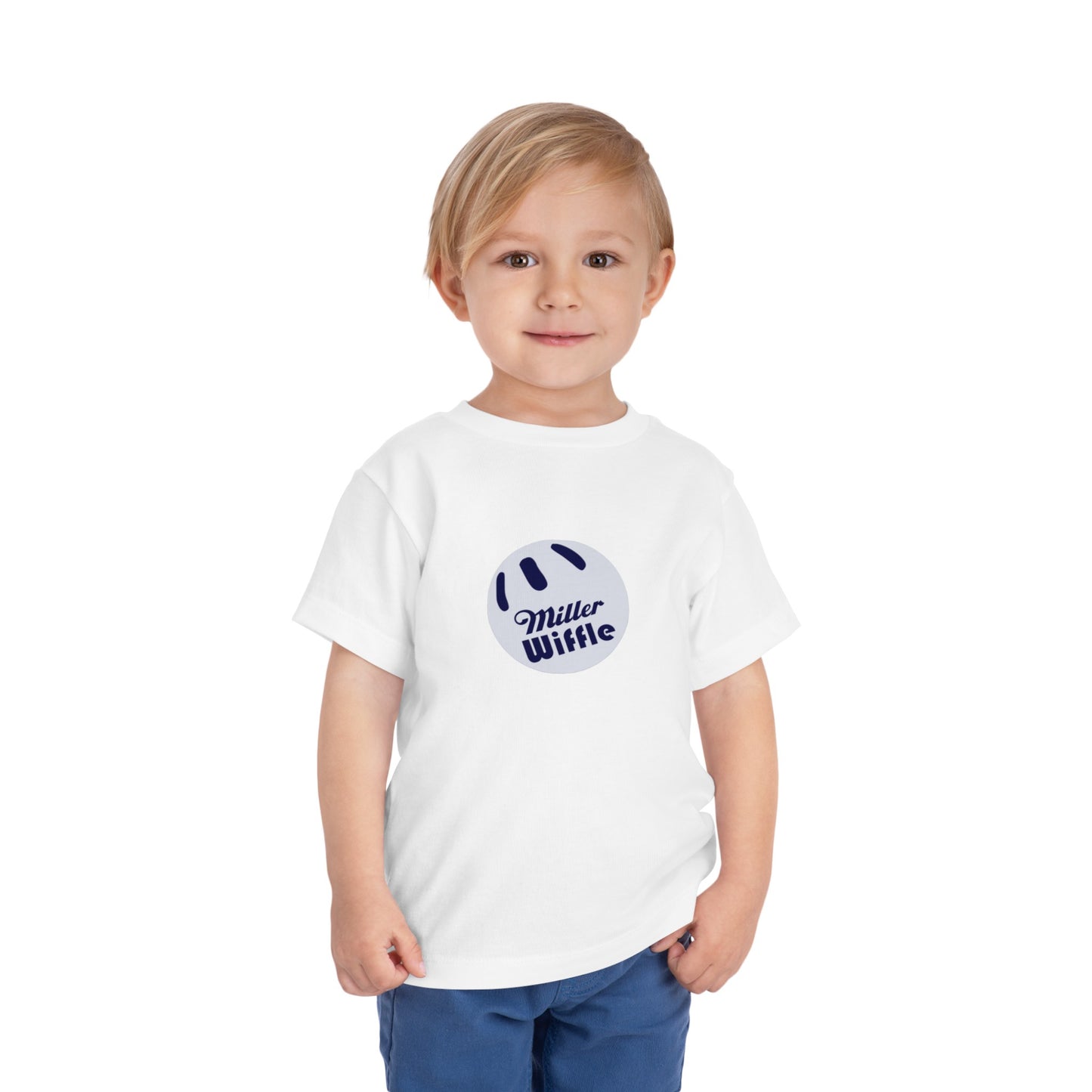 A young child with light hair stands against a plain white background, smiling while wearing a vibrant yellow Printify short sleeve tee named "Miller Wiffel Ball OG Logo - Toddler Short Sleeve Tee." The shirt features the "Miller Wiffle" text and a wiffle ball graphic on the front. The child is also dressed in blue pants.