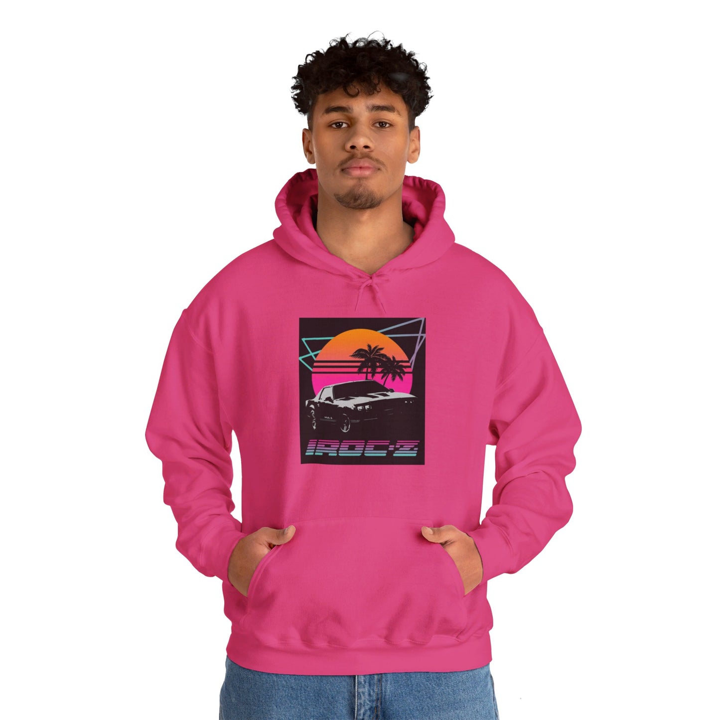 Introducing the Chevrolet IRoc Z28 - 1980s Retro Hoodie by Printify: This vibrant pink hoodie showcases a striking retro design on the front, featuring a classic American muscle car set against an orange and red sunset with palm trees, intersected by geometric shapes. The text "IROCZ" is prominently displayed below the image. Offering a relaxed fit and equipped with a front pocket, this hoodie perfectly captures the essence of 1980s style.