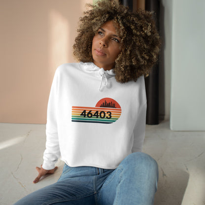 The Miller Beach 46403 Women's Crop Hoodie by Printify features a nostalgic design with a city skyline silhouette and the iconic "46403" zip code, set against a striped sunset backdrop in vibrant orange, green, and yellow hues that capture the essence of Miller Beach.