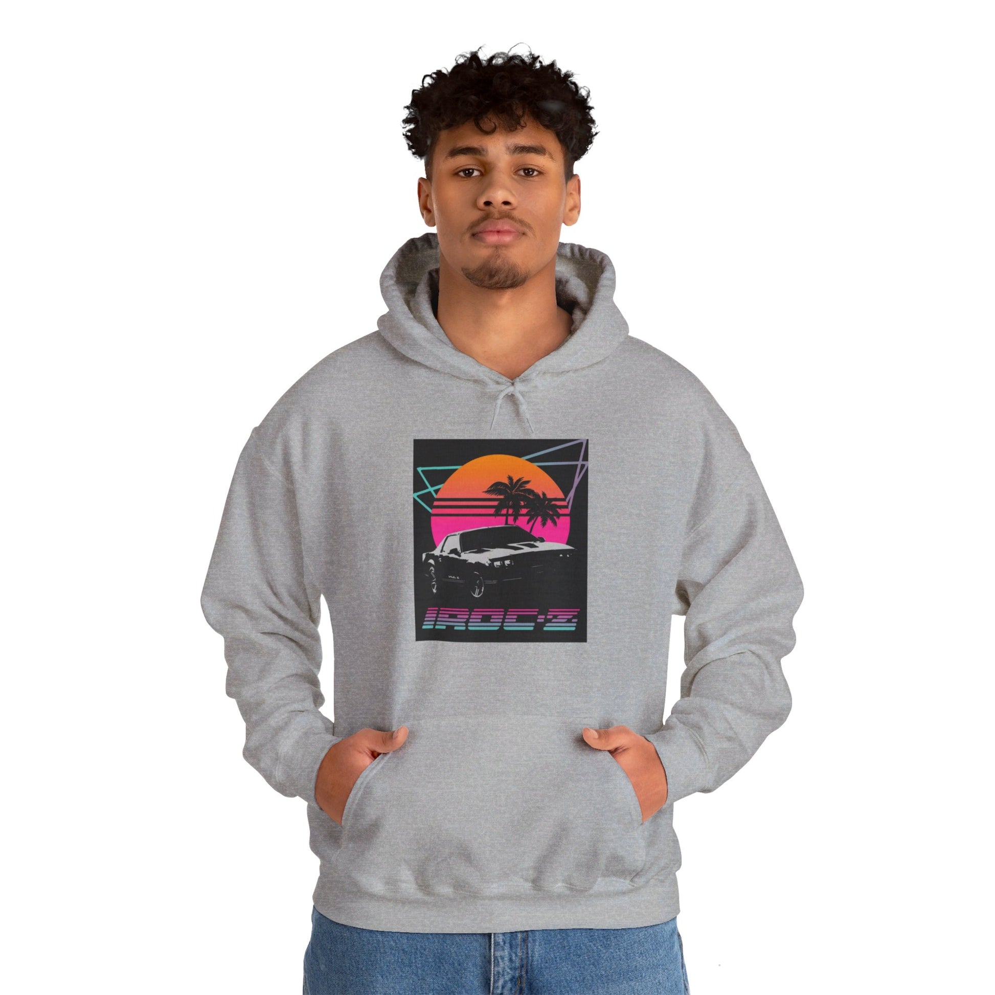 Introducing the Chevrolet IRoc Z28 - 1980s Retro Hoodie by Printify: This vibrant pink hoodie showcases a striking retro design on the front, featuring a classic American muscle car set against an orange and red sunset with palm trees, intersected by geometric shapes. The text "IROCZ" is prominently displayed below the image. Offering a relaxed fit and equipped with a front pocket, this hoodie perfectly captures the essence of 1980s style.