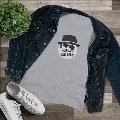 A cozy red t-shirt, the Miller Wiffel Ball - Blues Brothers - Women's Triblend Tee by Printify, features a graphic of a masked face wearing a fedora hat and sunglasses, along with the text "Miller Wiffle." The t-shirt is displayed on a wooden floor, paired with a denim jacket, white sneakers, and a potted green plant to create a vintage look.