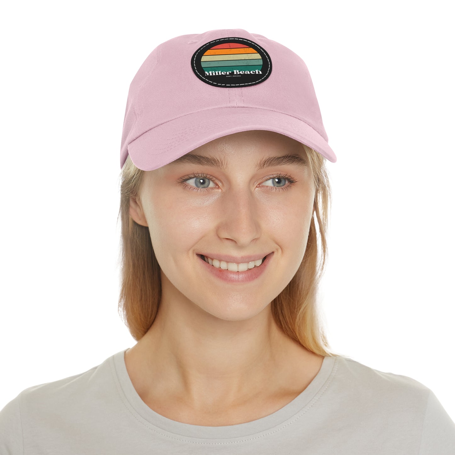 The Miller Beach Retro Sunset - Dad Hat with Leather Patch (Round) by Printify is a pink baseball cap crafted from bio-washed chino twill for added comfort. It features a PU leather patch adorned with horizontal stripes in red, orange, yellow, green, and blue. Below the stripes, "Miller Beach" is embroidered in white. An adjustable strap at the back ensures a perfect fit.