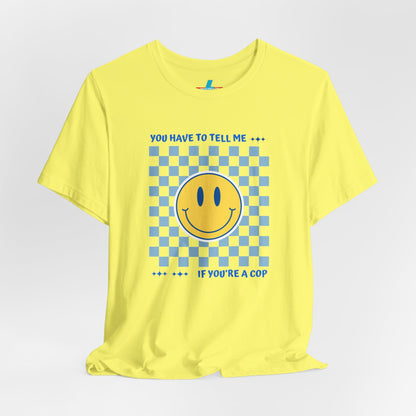 A light yellow unisex jersey short sleeve tee from Printify featuring a blue and yellow checkerboard pattern with a large yellow smiley face in the center. The shirt displays blue text above and below the pattern that reads, "You have to tell me if you're a cop." It's made from 100% Airlume combed and ring-spun cotton.