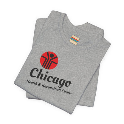 Two gray T-shirts are neatly folded on top of each other, with the top shirt showcasing the text "Chicago Health & Racquetball Clubs" in black below a red logo featuring a person with raised arms inside a circular design. This retro-inspired tee, named "Chicago Health Clubs 1980s Retro - Unisex Jersey Short Sleeve Tee" by Printify, is perfect for any fan of Chicago Health Clubs.