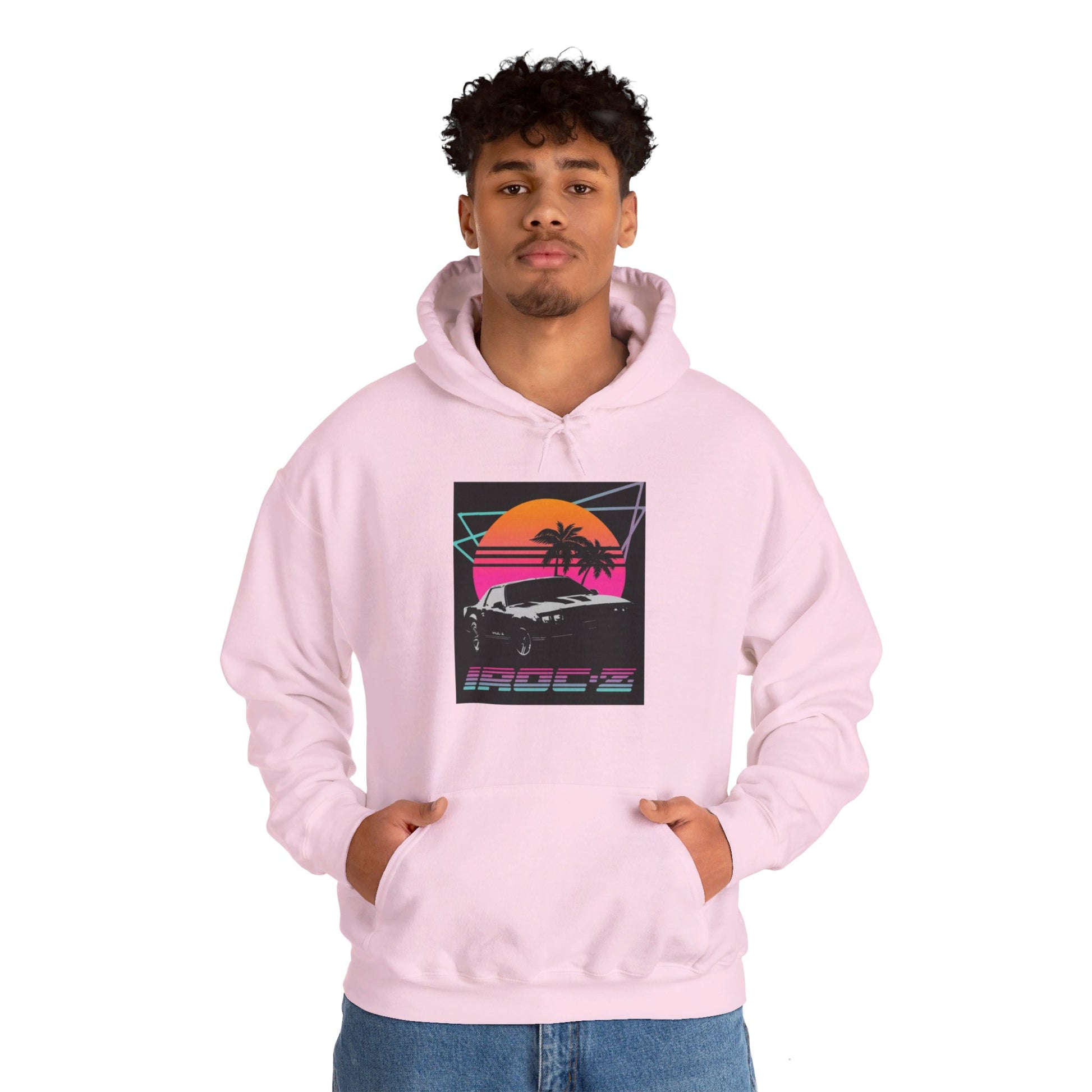 Introducing the Chevrolet IRoc Z28 - 1980s Retro Hoodie by Printify: This vibrant pink hoodie showcases a striking retro design on the front, featuring a classic American muscle car set against an orange and red sunset with palm trees, intersected by geometric shapes. The text "IROCZ" is prominently displayed below the image. Offering a relaxed fit and equipped with a front pocket, this hoodie perfectly captures the essence of 1980s style.