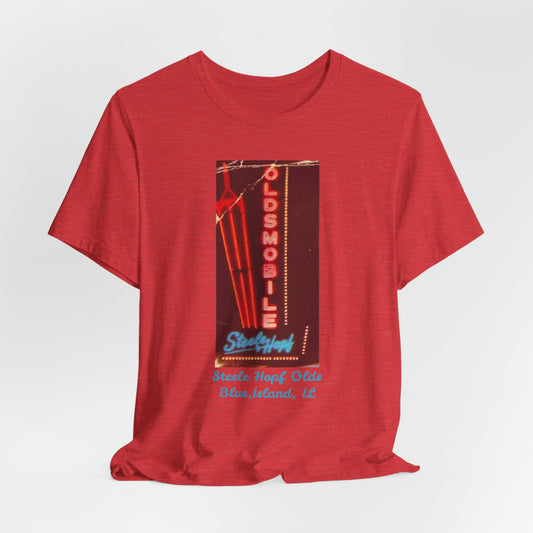 The Blue Island Illinois Steele-Hopf Oldsmobile Unisex Jersey Short Sleeve Tee by Printify features a vibrant red design with an image of a vintage neon sign. The sign displays "OLDSMOBILE" in vertical red letters and "Strafford" in blue cursive script. Below the sign, light blue text reads "Sleets Hopf Olds" and "Blue Island, IL," making it an ideal retro-inspired tee for classic car enthusiasts.