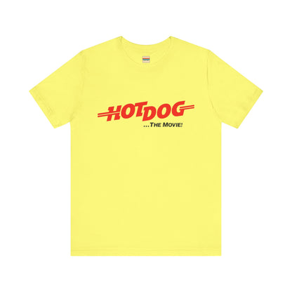 A smiling man and woman interact while wearing matching yellow "Hot Dog The Movie 1984 - Unisex Jersey Short Sleeve Tee" by Printify, featuring bold red "HOT DOG THE MOVIE" text. Their vibrant tees perfectly complement their blue jeans, with the man casually having his hands in his pockets and the woman resting one hand on his shoulder.