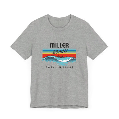 The Miller Beach 46403 Surf Style - Unisex Jersey Short Sleeve Tee by Printify features a retro surf-inspired design on a yellow background. The shirt displays "MILLER BEACH" above waves with a skyline illustration, while "Cary, IN 46403" is printed below the waves. The vibrant design also includes colorful stripes in shades of blue, red, and orange.