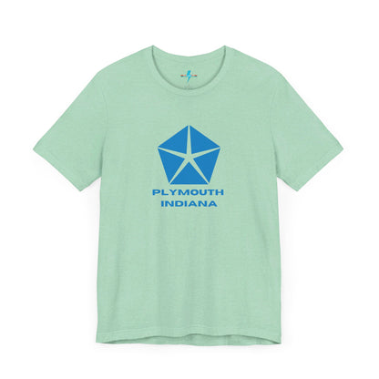 A black Printify Plymouth, Indiana Chrysler Logo T-shirt is displayed against a plain white background. The shirt features a blue, five-point star emblem reminiscent of a vintage autos design above the text "PLYMOUTH INDIANA" in blue, centered across the front. The shirt is laid out flat with the sleeves slightly bent.