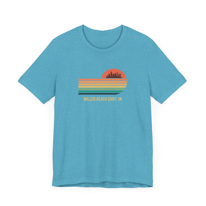 Introducing the Miller Beach Gary, IN Sunset Gradient - Unisex Jersey Short Sleeve Tee by Printify. This stylish blue T-shirt features a circular sunset graphic on the chest, showcasing a black city skyline silhouette against an orange-red sunset with horizontal stripes in green, yellow, and orange. Below the graphic reads "MILLER BEACH GARY, IN." Crafted from soft cotton for extra beachy charm.