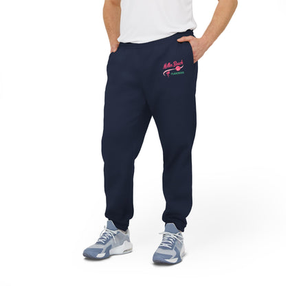 A person is confidently dressed in the cozy Miller Beach Flamingos - Adidas Unisex Fleece Joggers from Printify, featuring "Myrtle Beach" and "Yearround" text in pink and green on the left thigh. They complete their look with a white shirt and gray sneakers, standing against a plain white background.