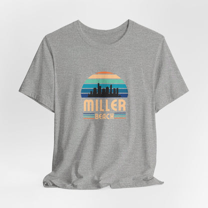 The Printify Miller Beach Chicago Skyline - Unisex Jersey Short Sleeve Tee is a light blue T-shirt featuring a stylized graphic of the Chicago skyline against a setting sun with gradient shades of blue and orange. Below the graphic, "Miller Beach" is printed in bold, yellow letters.