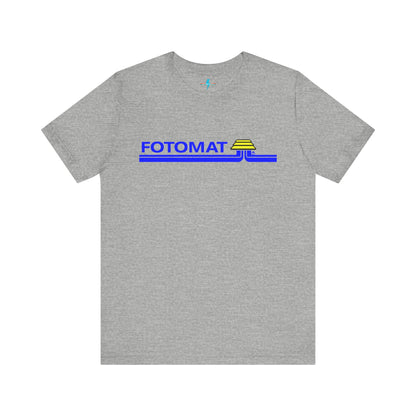 The Fotomat 1970s Retro T-Shirt by Printify is a dark gray short-sleeve shirt that features the word "FOTOMAT" in bold blue capital letters and a blue and yellow graphic design resembling layered lines and an upside-down triangle. This Tshirt captures the essence of 80s nostalgia with its centered, retro design.