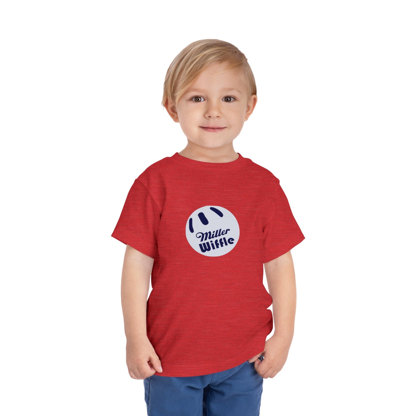 A young child with light hair stands against a plain white background, smiling while wearing a vibrant yellow Printify short sleeve tee named "Miller Wiffel Ball OG Logo - Toddler Short Sleeve Tee." The shirt features the "Miller Wiffle" text and a wiffle ball graphic on the front. The child is also dressed in blue pants.