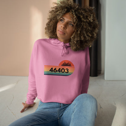 The Miller Beach 46403 Women's Crop Hoodie by Printify features a nostalgic design with a city skyline silhouette and the iconic "46403" zip code, set against a striped sunset backdrop in vibrant orange, green, and yellow hues that capture the essence of Miller Beach.