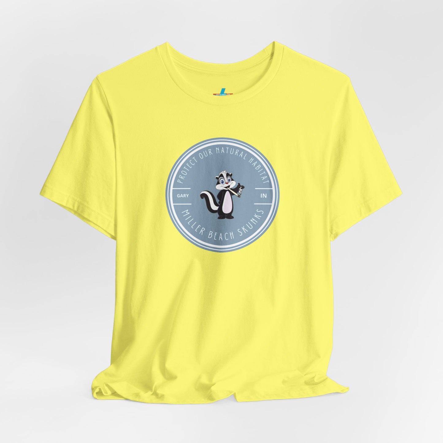 The Printify Miller Beach Skunks - Unisex Jersey Short Sleeve Tee is a gray T-shirt that features a circular blue logo at the center. Inside the logo, there is an illustration of a skunk with the text "Protect Our Habitat" and "Miller Beach Skunks" around the border, promoting environmental consciousness. The upper left part of the logo reads "Gary, IN.