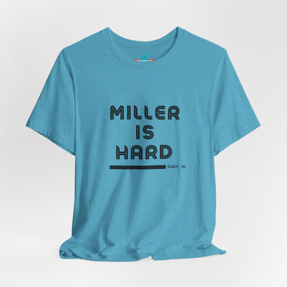 A unisex jersey short sleeve tee from Printify in burnt orange features the bold black text "MILLER IS HARD" on the chest, with "GARY, IN" written beneath in smaller black font. Perfect for showcasing Miller Beach's iconic spirit, this t-shirt is displayed against a plain white background.