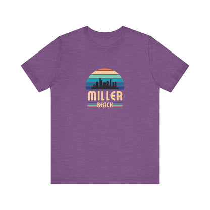 The Printify Miller Beach Chicago Skyline - Unisex Jersey Short Sleeve Tee is a light blue T-shirt featuring a stylized graphic of the Chicago skyline against a setting sun with gradient shades of blue and orange. Below the graphic, "Miller Beach" is printed in bold, yellow letters.