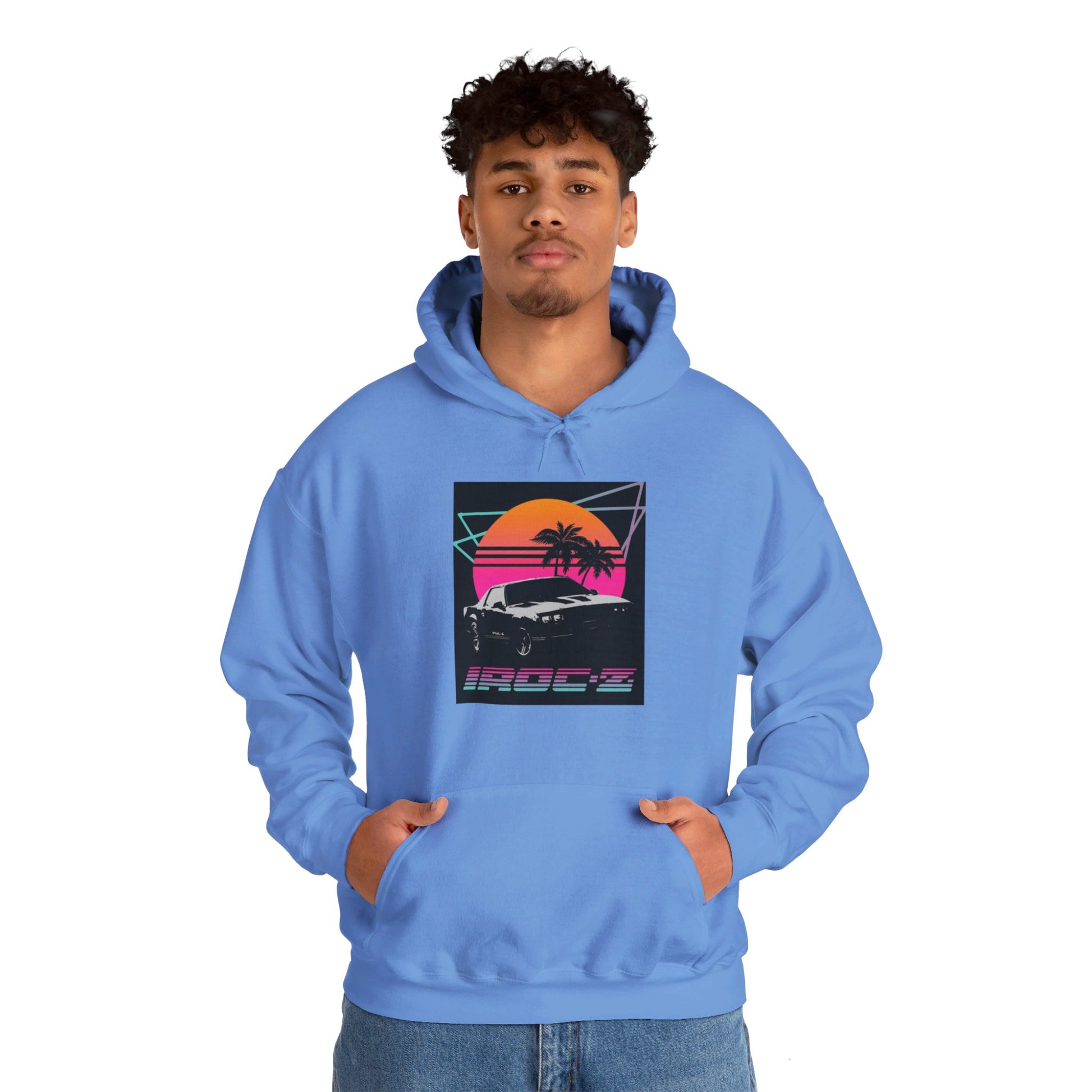 Introducing the Chevrolet IRoc Z28 - 1980s Retro Hoodie by Printify: This vibrant pink hoodie showcases a striking retro design on the front, featuring a classic American muscle car set against an orange and red sunset with palm trees, intersected by geometric shapes. The text "IROCZ" is prominently displayed below the image. Offering a relaxed fit and equipped with a front pocket, this hoodie perfectly captures the essence of 1980s style.