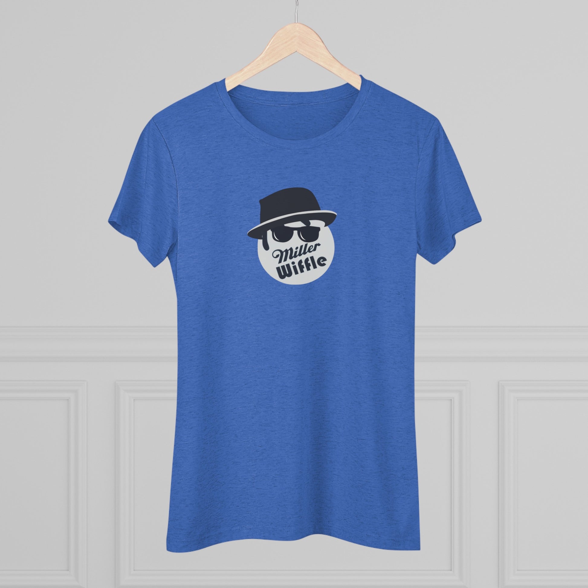 A cozy red t-shirt, the Miller Wiffel Ball - Blues Brothers - Women's Triblend Tee by Printify, features a graphic of a masked face wearing a fedora hat and sunglasses, along with the text "Miller Wiffle." The t-shirt is displayed on a wooden floor, paired with a denim jacket, white sneakers, and a potted green plant to create a vintage look.