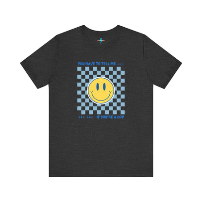 A light yellow unisex jersey short sleeve tee from Printify featuring a blue and yellow checkerboard pattern with a large yellow smiley face in the center. The shirt displays blue text above and below the pattern that reads, "You have to tell me if you're a cop." It's made from 100% Airlume combed and ring-spun cotton.