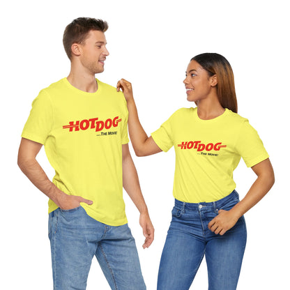 A smiling man and woman interact while wearing matching yellow "Hot Dog The Movie 1984 - Unisex Jersey Short Sleeve Tee" by Printify, featuring bold red "HOT DOG THE MOVIE" text. Their vibrant tees perfectly complement their blue jeans, with the man casually having his hands in his pockets and the woman resting one hand on his shoulder.