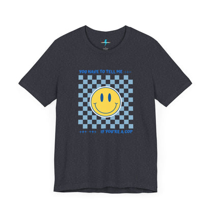 A light yellow unisex jersey short sleeve tee from Printify featuring a blue and yellow checkerboard pattern with a large yellow smiley face in the center. The shirt displays blue text above and below the pattern that reads, "You have to tell me if you're a cop." It's made from 100% Airlume combed and ring-spun cotton.