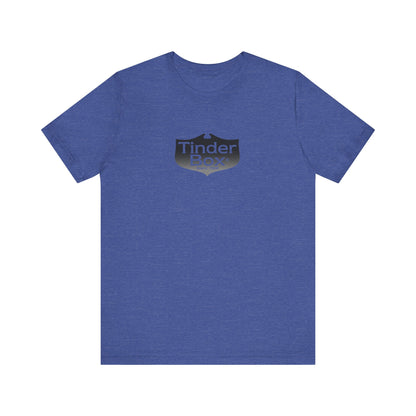 A light blue Printify "The Tinder Box 1980s" unisex jersey short sleeve tee featuring the "Tinder Box" logo in the center. The logo consists of a dark shield with the words "Tinder Box" in white text, with a small flame icon replacing the dot above the "i".