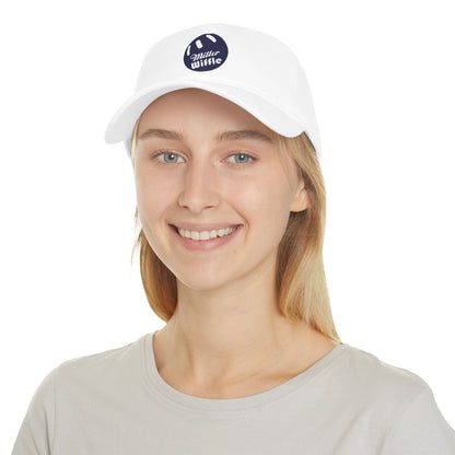 A person with long blonde hair smiles while wearing the Miller Beach Wiffel Ball - Low Profile Baseball Cap by Printify. The cap, crafted from 100% cotton, is white with a navy blue circular logo that reads "Miller Wiffle." The individual is also dressed in a light grey t-shirt, set against a plain white background.