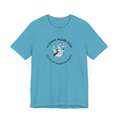 A unisex jersey short sleeve tee in orange from Printify, known as the "Tot Lot Wizard - Miller Beach," features a central graphic of a wizard holding a wand. The design is surrounded by stars and lightning bolts and includes the text "I'M DOING WIZARD STUFF" and "THE TOT LOT WIZARD MILLER BEACH.