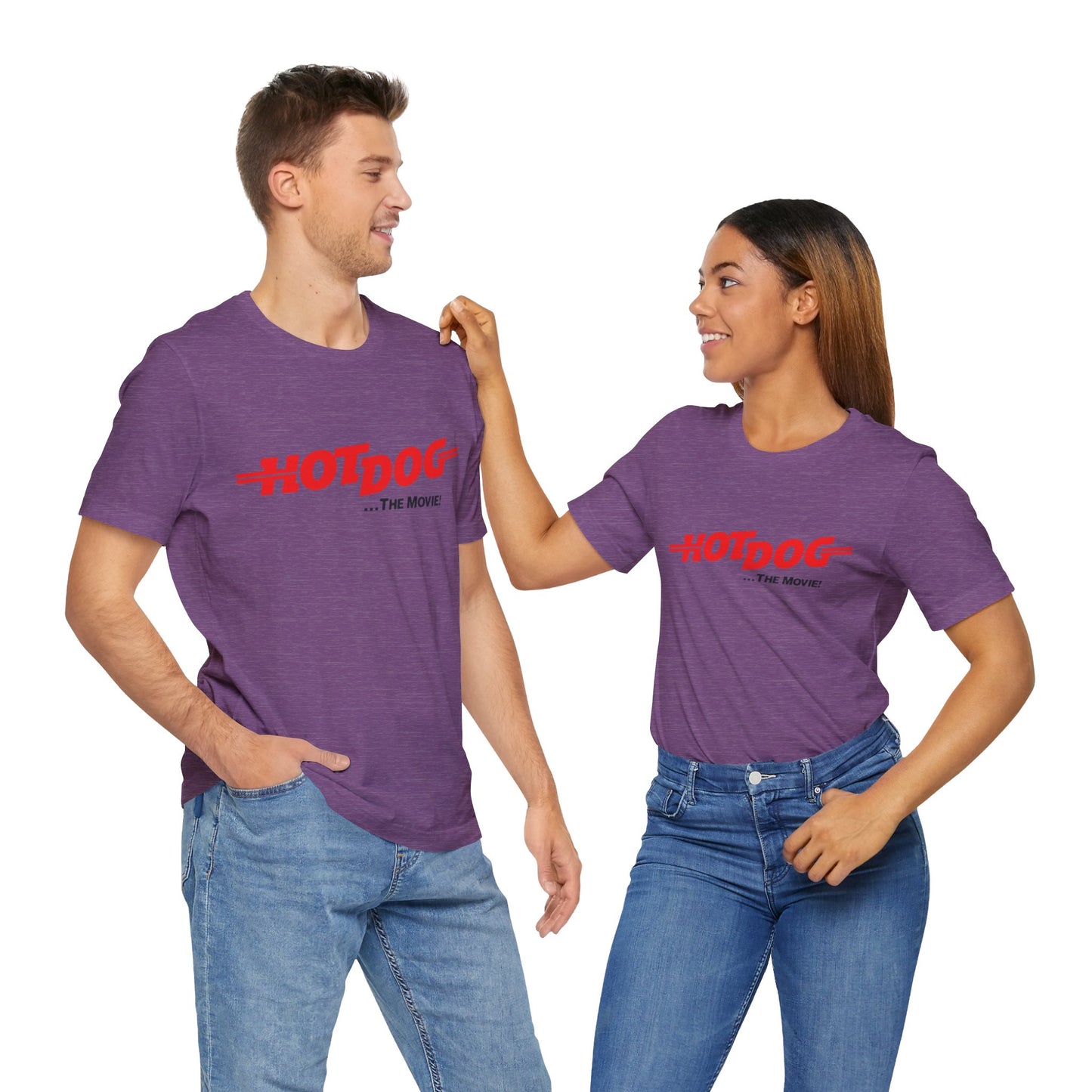 A smiling man and woman interact while wearing matching yellow "Hot Dog The Movie 1984 - Unisex Jersey Short Sleeve Tee" by Printify, featuring bold red "HOT DOG THE MOVIE" text. Their vibrant tees perfectly complement their blue jeans, with the man casually having his hands in his pockets and the woman resting one hand on his shoulder.