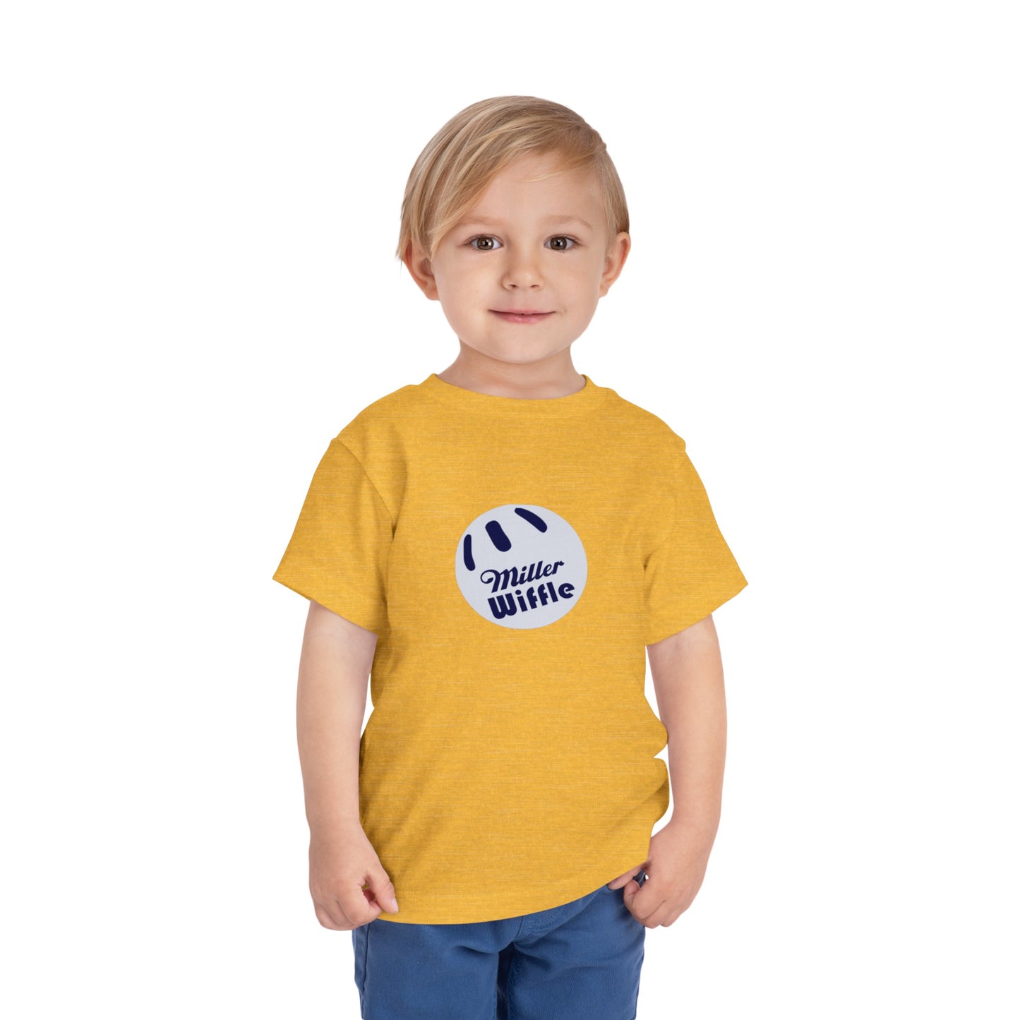 A young child with light hair stands against a plain white background, smiling while wearing a vibrant yellow Printify short sleeve tee named "Miller Wiffel Ball OG Logo - Toddler Short Sleeve Tee." The shirt features the "Miller Wiffle" text and a wiffle ball graphic on the front. The child is also dressed in blue pants.