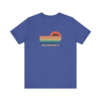 Introducing the Miller Beach Gary, IN Sunset Gradient - Unisex Jersey Short Sleeve Tee by Printify. This stylish blue T-shirt features a circular sunset graphic on the chest, showcasing a black city skyline silhouette against an orange-red sunset with horizontal stripes in green, yellow, and orange. Below the graphic reads "MILLER BEACH GARY, IN." Crafted from soft cotton for extra beachy charm.