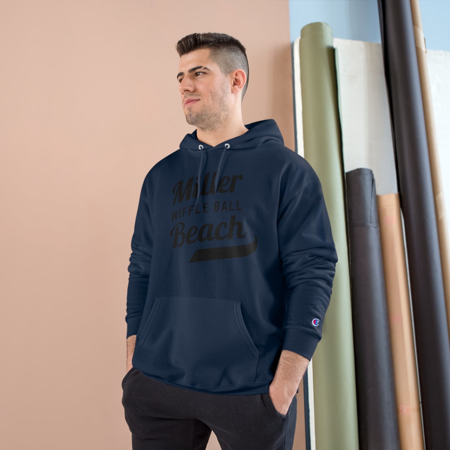 A man wearing a Champion Hoodie from Printify, featuring the "Miller Beach Wiffel Ball Retro Jersey Logo," stands against a peach-colored wall and vertical rolls of various colored fabrics. He has short, dark hair and is looking slightly to the left with a relaxed expression.