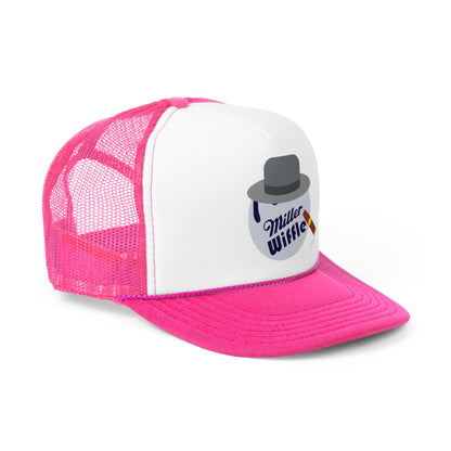 A smiling person with long hair and a nose ring wears the Miller Wiffel Ball ITYSL Driving Crooner Trucker Cap from Printify, which is white and pink and features a graphic of a hat, pipe, and "Miller Wiffle" text. The cap includes an adjustable snap closure for comfort. They are also wearing a blue denim jacket over a white shirt.