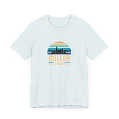 The Printify Miller Beach Chicago Skyline - Unisex Jersey Short Sleeve Tee is a light blue T-shirt featuring a stylized graphic of the Chicago skyline against a setting sun with gradient shades of blue and orange. Below the graphic, "Miller Beach" is printed in bold, yellow letters.