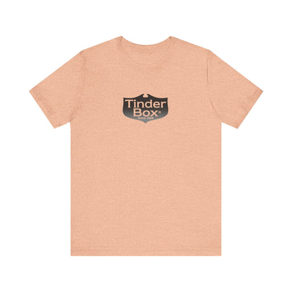 A light blue Printify "The Tinder Box 1980s" unisex jersey short sleeve tee featuring the "Tinder Box" logo in the center. The logo consists of a dark shield with the words "Tinder Box" in white text, with a small flame icon replacing the dot above the "i".
