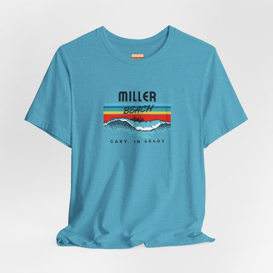 The Miller Beach Surf Style - Unisex Jersey Short Sleeve Tee by Printify comes in light blue, showcasing a vibrant design featuring waves and a sunset with the text "Miller Beach" above it. Below the image, the words "Gary, IN 46403" are printed, highlighting this unique offering from a small local business. The design also includes striking stripes in red, yellow, and blue.