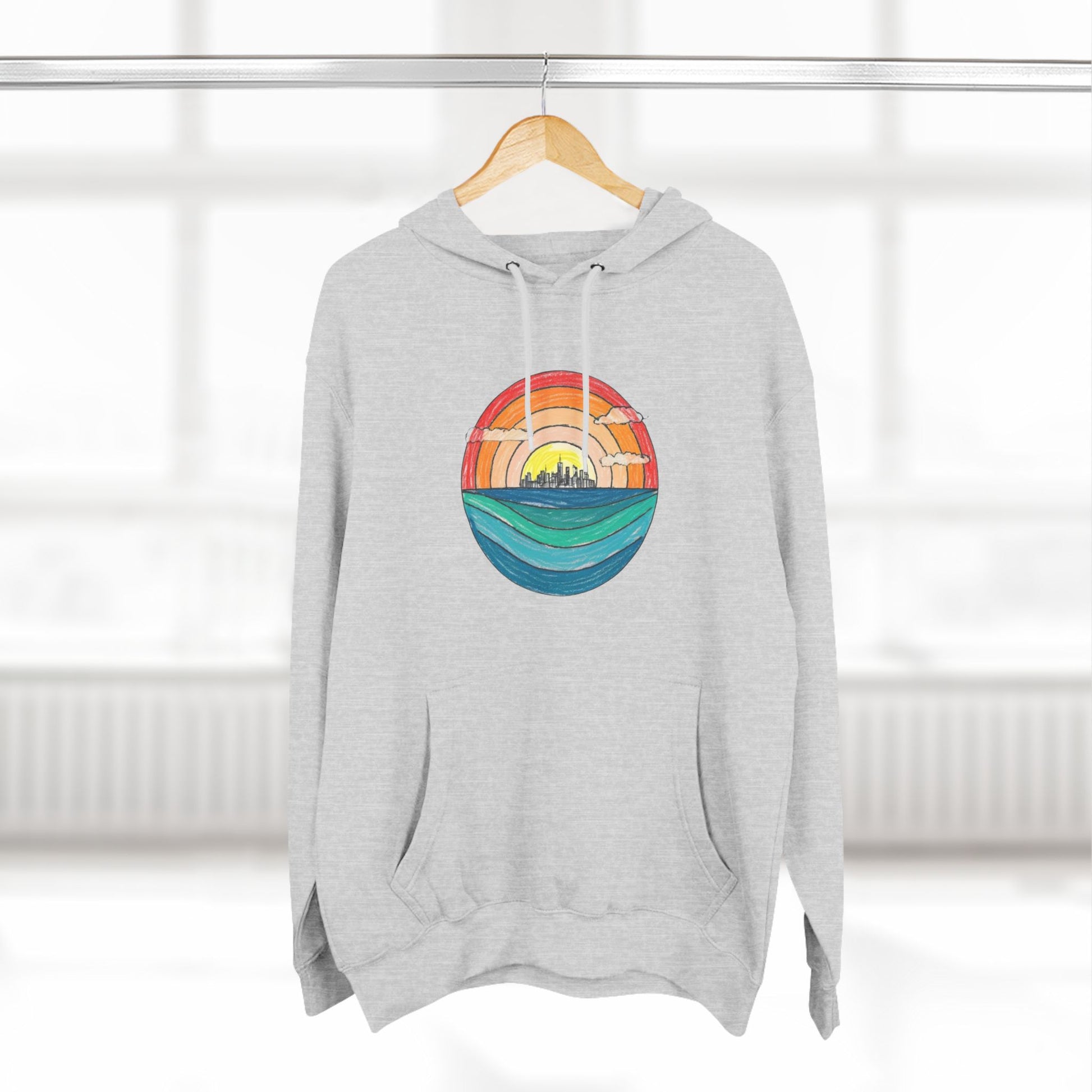 The Miller Beach Abstract - Three-Panel Fleece Hoodie by Printify is a premium light gray pullover featuring a vibrant circular front design. It showcases an abstract sunset-over-ocean scene with blue waters, an orange to red gradient sky, and a city skyline silhouette. Made from soft combed ring-spun cotton and lined with cozy fleece for extra warmth.