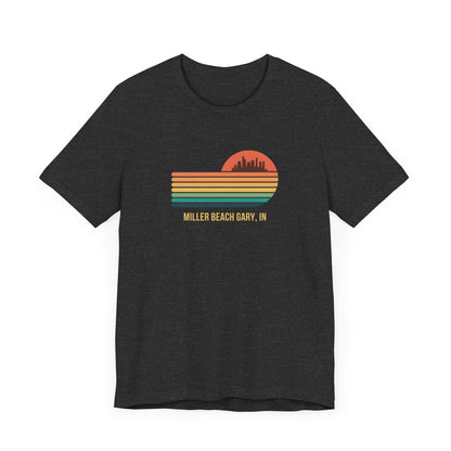 Introducing the Miller Beach Gary, IN Sunset Gradient - Unisex Jersey Short Sleeve Tee by Printify. This stylish blue T-shirt features a circular sunset graphic on the chest, showcasing a black city skyline silhouette against an orange-red sunset with horizontal stripes in green, yellow, and orange. Below the graphic reads "MILLER BEACH GARY, IN." Crafted from soft cotton for extra beachy charm.