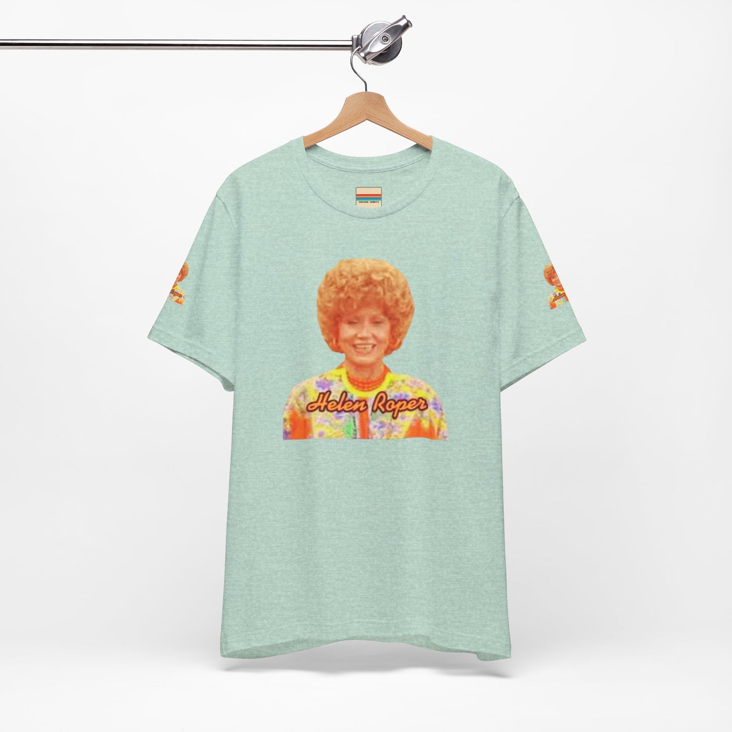 A lavender-colored Helen (Mrs.) Roper Three's Company - Unisex Jersey Short Sleeve Tee by Printify is hanging on a wooden hanger against a white background. This T-shirt features an image of a smiling person with curly hair wearing a colorful blouse, with the text "Helen Roper" underneath the image. Both sleeves have identical designs, making it perfect Three's Company merchandise.