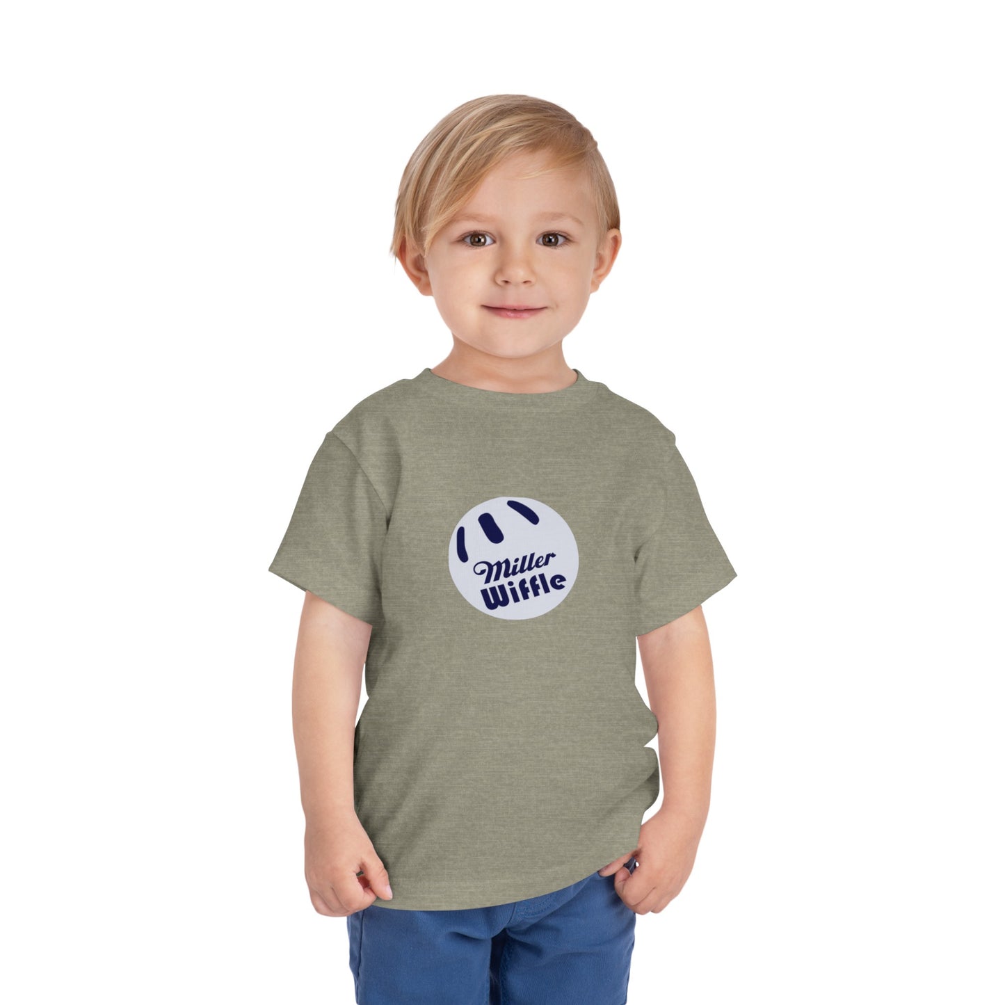 A young child with light hair stands against a plain white background, smiling while wearing a vibrant yellow Printify short sleeve tee named "Miller Wiffel Ball OG Logo - Toddler Short Sleeve Tee." The shirt features the "Miller Wiffle" text and a wiffle ball graphic on the front. The child is also dressed in blue pants.