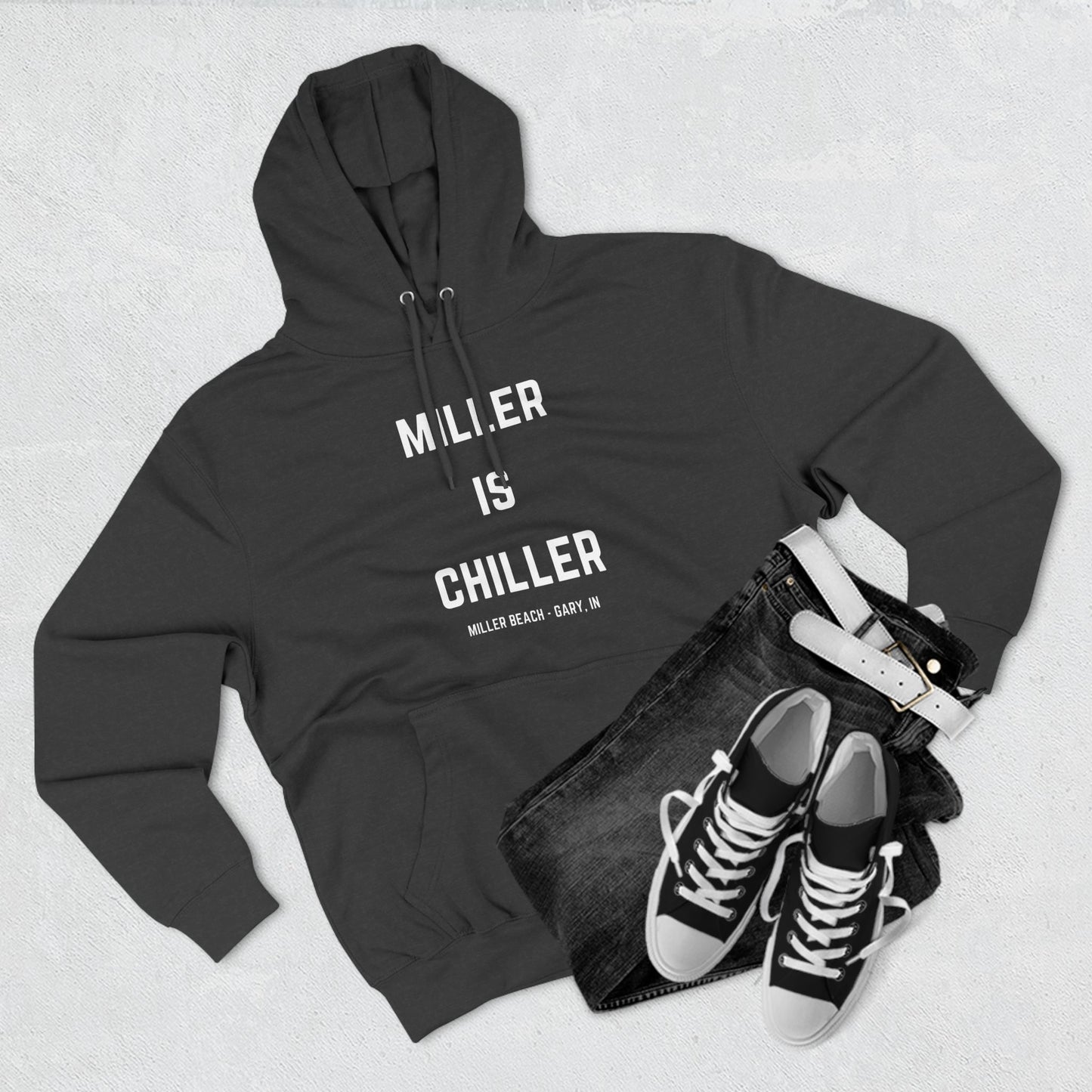 The Miller is Chiller - Miller Beach Three-Panel Fleece Hoodie from Printify features bold white text reading "MILLER IS CHILLER" on the front, with smaller text below stating "MILLER BEACH - GARY, IN." This comfortable black hoodie includes a front pocket and drawstrings.