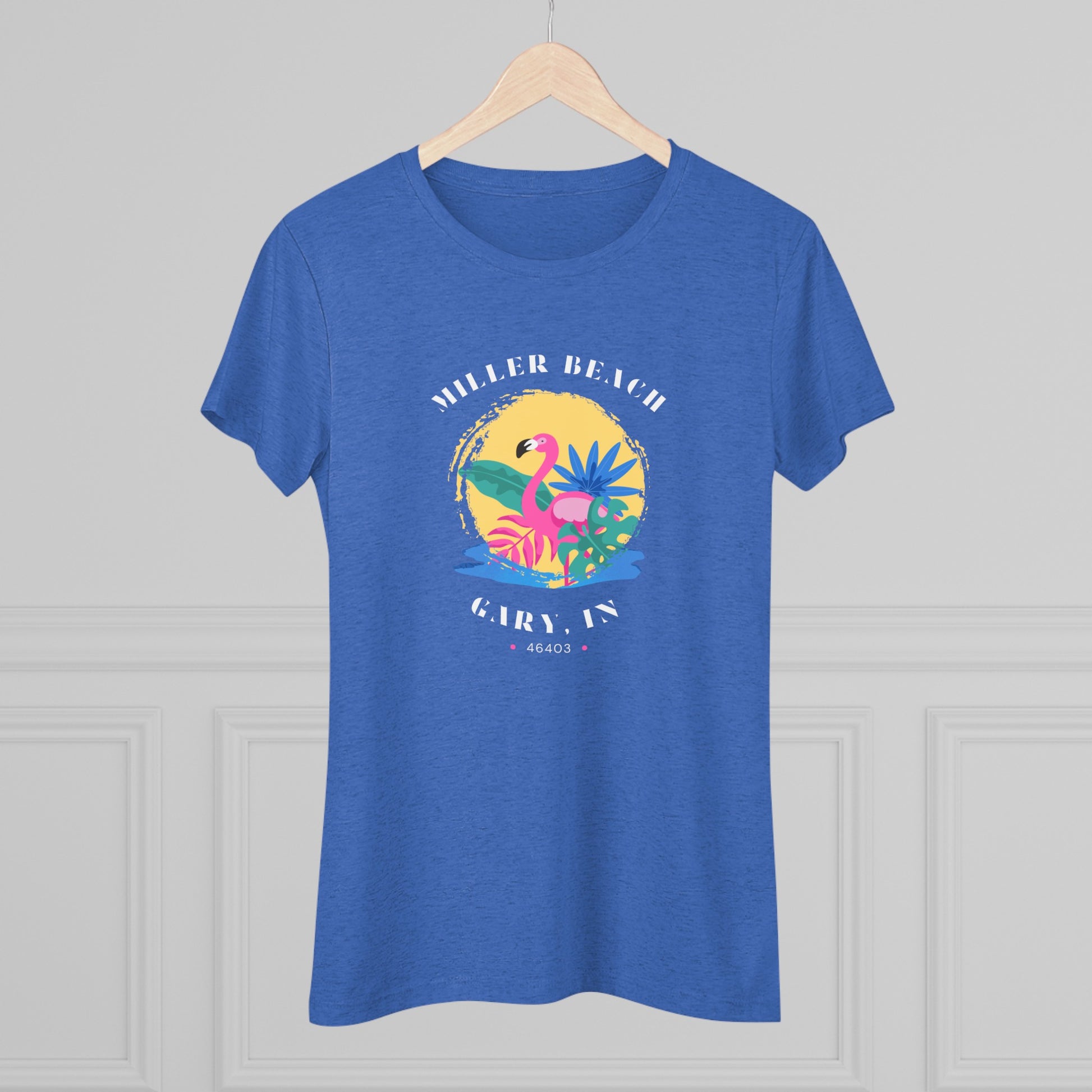 A women's triblend tee from Printify, the Miller Beach Flamingo features a blue color and "Miller Beach Gary, IN 46403" text with a vibrant design of a flamingo amidst tropical foliage for a vintage aesthetic. This t-shirt is showcased on a wooden surface alongside a denim jacket, white sneakers, and a potted plant.